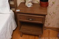 Photo of first night table showing wood veneer top, drawer with silver metal handle, open shelf, and four legs next to bed.
