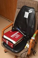 Black Homedics back massager seat pad placed on brown cushioned wooden armchair with books, showing massager and remote control cable.