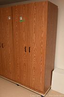 Front view of a tall faux wood armoire cabinet with two doors and two black handles, showing the wood grain pattern and stickers on the upper left door.