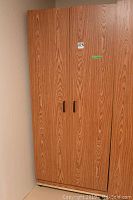 Full front view of tall faux wood armoire cabinet with two doors and black handles against corner wall.