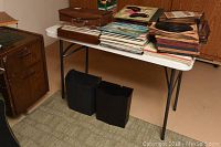 White plastic folding table with metal legs, shown with magazines and two black bins underneath.