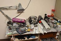 Wide view showing assembled tools, hardware, and other objects on tabletop including power saws, clamps, extension cords, small parts, and work lamp.