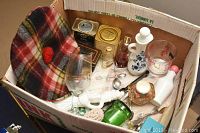 Box containing a variety of collectible and household items including bottles, tins, a glass wine glass, porcelain bottle, baby brush, and plaid fabric item.