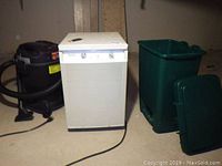 Three items next to each other on concrete floor: black/red shop vac, white Danby dehumidifier with knobs, green plastic garbage can with lid off.
