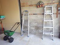 Full view of items in garage: 5-foot aluminum ladder, 2-foot platform aluminum ladder, green Scotts Speedy Green 1000 spreader, faux flowers on wall