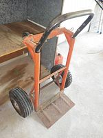 Photo showing the orange metal utility cart from a front angle, revealing rust on the base platform and wear on the frame and handle.