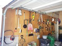 Wide shot of garage wall with various garden and hand tools hanging on pegboard including shovels, pruners, rakes, and wheelbarrow on the floor