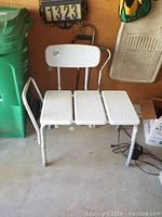 White adjustable bath seat with perforated seat panels and backrest, placed on a garage floor next to a green recycle bin and a garage wall with a house number plate above.