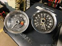 Photo showing front view of the Jaeger tachometer (left) and speedometer (right) gauges resting on a black surface with dial faces and needles visible.