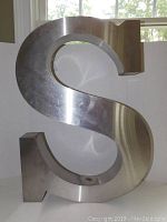 Front and side views of a large metal sculpture shaped like the letter S, showing brushed metal surface and block serif font style.