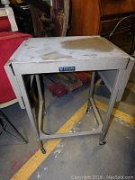 Side view of vintage gray metal typewriter cart showing rectangular top and frame with caster wheels.