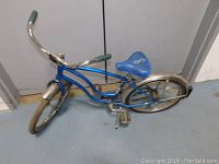 Side view of blue child's bicycle showing frame, blue saddle with logo, hand grips, and rear chrome fender.