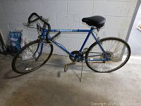 Blue road bicycle leaning on kickstand against a concrete wall with visible U.S. General Coach branding on frame.