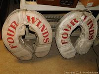 Pair of white horseshoe/vintage style life jackets with red "FOUR WINDS" lettering, shown side by side on floor.