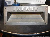 Front view of rectangular metal slot with 'PAPERS' embossed on top, showing some usage wear and dirt.