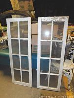 Pair of vintage wooden framed windows, both standing upright against a background with boxes, showing eight glass panes each and white painted frames with wear and chipping.