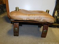 Front view of vintage leather saddle stool showing leather seat, wooden legs, and brass hardware.