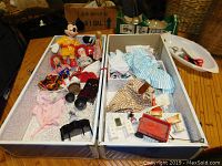 Two open compartments of a chest holding dolls, doll clothes, and accessories spread out