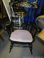 Black wooden rocking chair with painted floral stencil details and pink cushion, shown from front angle with some surrounding furniture visible.