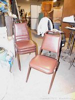 Four brown vinyl padded metal stacking chairs, two stacked, two unstacked in cluttered room showing condition.