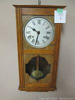 Front view of solid oak antique box wall clock showing Roman numeral dial and visible pendulum behind glass panel.