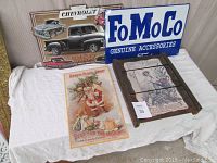 Four vintage advertising signs on a white cloth: Chevrolet Chevy Trucks metal sign, blue and white FoMoCo Ford sign, wooden Burpee seed sign, vintage Santa Claus soap sign.