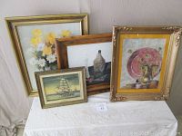 Four framed paintings arranged on a table against a wall: one featuring yellow and white flowers, one with a sailing ship, another with bottles, and a large still life with a pink plate and brass pitcher.