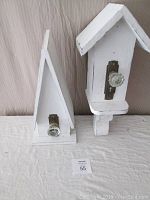 Two white Victorian-style handcrafted birdhouses side by side against beige fabric background, showing one intended for sitting and the other for hanging, each with glass antiques knobs on front.