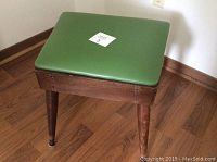 Side and partial top view of wooden stool showing stained brown finish, tapered wooden legs, and upholstery material on seat.