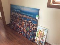 Two New York themed artworks leaning against a wall; large cityscape print and smaller colorful sketch.