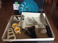 Overview of entire kitchen utensil lot displayed with a blue colander in the background and various utensils in a cardboard tray including knife, wine opener, measuring spoons, egg cups, spaghetti measurer, sieve and ice cream scoop