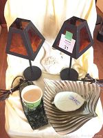 Photo showing two black iron bedside lamps with square glass-inset lampshades, large fluted silver bowl, leaf-shaped glass dish with dragonfly, rectangular green tray with small cup, and wall-hung half pitcher piece in the background