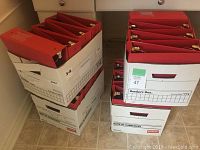 Photo showing 26 red binders stored upright in white banker's boxes on a tiled floor.