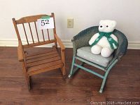 Photo of two antique children's rocking chairs and a teddy bear on wooden floor. Wooden rocker chair is natural wood with vertical slats on back and slatted seat. Wicker rocker is painted greenish-blue with woven wicker construction, cushion seat, and white teddy bear with green scarf sitting on it.