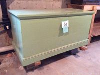 Front angled view of a large light green painted solid wood storage chest with hinged lid and a label on the front.