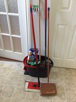 Front view showing mop with red handle, bucket with red pedal-operated spinner, Mr. Clean Eraser box, mop pads, and carpet sweeper leaning against a wall