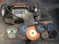 Photo showing the Delta 6 inch bench grinder with the transparent safety shield and the power cord. Below the grinder are nine grinding disks of various types and colors arranged in a row.