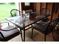 Full view of patio table with glass top and four cast aluminum chairs with cushions in an indoor setting.