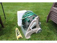 Photo of beige plastic hose reel with green garden hose coiled on it, featuring a hand crank and wheels, set on grass with nearby stacked brown chairs.