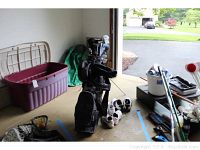 Golf bag filled with golf clubs standing upright in garage along with three pairs of golf shoes and a large plastic bin to the side.