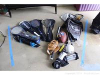 Wide shot including rackets, ball glove, football, badminton set, lacrosse sticks, bat, volleyball, and bags on concrete floor.
