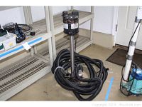 Full view of sump pump with coiled discharge hose on floor, showing motor housing and metal shaft.