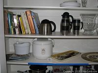 Cookbooks, rice cooker, salad spinner, serving platters and kitchenware arranged on white shelves.