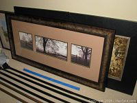 Three framed wall arts stacked on floor: a large decorative triptych with tree and fence images, a smaller decorative triptych with similar theme, and a framed watercolor painting of the Sultan Mosque.