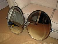 Pair of oval wall-mount tilt mirrors showing chrome frames and reflective glass surfaces laying on carpeted floor in front of sofa.
