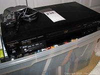 Panasonic black DVD player on top of plastic storage container holding DVDs, remote on top with power cable coiled
