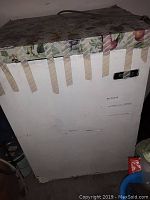 Small white freezer with floral adhesive top and taped edges, showing signs of wear and scuffs.