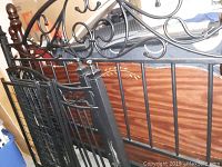 Photo showing black metal ornate king size bed headboard and black metal bed frame pieces leaning against a wooden furniture piece with floral inlay design.