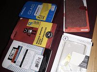 Photo showing six phone accessories including two tempered glass screen protectors (Mobest and PRO brands), one reddish fabric folio phone case, and clear protective phone cases for Samsung Galaxy.