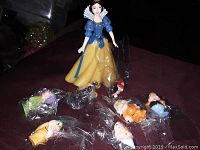 Photo displaying Snow White alongside the individually bagged dwarf figurines arranged on a dark surface.